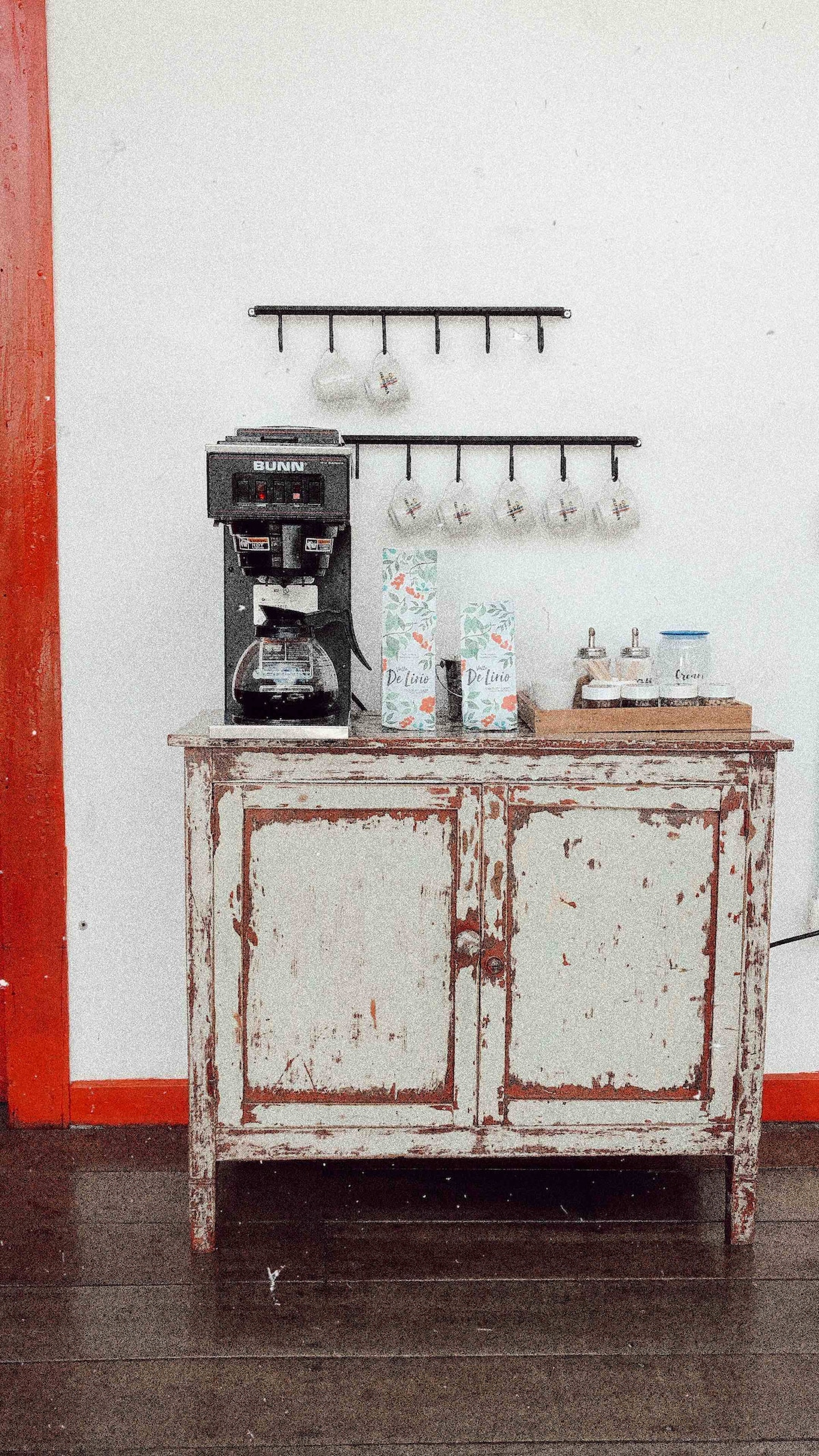 A vintage-style coffee station is set on a rustic wooden cabinet, featuring a modern coffee maker and a selection of coffee accessories. Mugs are neatly hung on a rack above, while a small assortment of cups and creamers is displayed on the cabinet's surface.