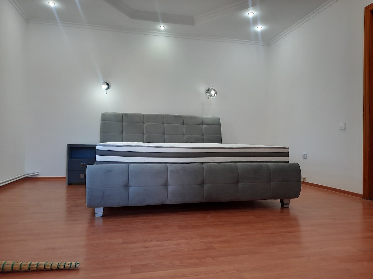 A spacious bed with a modern upholstered frame is centered against a neutral wall. Soft lighting fixtures are mounted above, providing an even glow. A wooden drawer unit is positioned beside the bed, enhancing the minimalist aesthetic of the room with warm-toned wooden flooring.