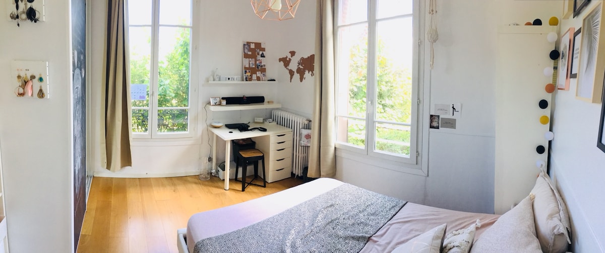 A bright bedroom features a spacious double bed dressed in soft linens. A desk area with a chair is positioned near large windows, providing natural light and views of the garden. Decorative elements include a world map and string lights, adding a welcoming touch.