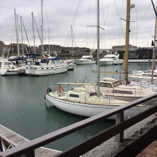A view of a marina filled with various boats docked in calm waters. The image captures multiple sailboats and yachts, with buildings visible in the background. A wooden railing is seen in the foreground, framing the scenic waterway.