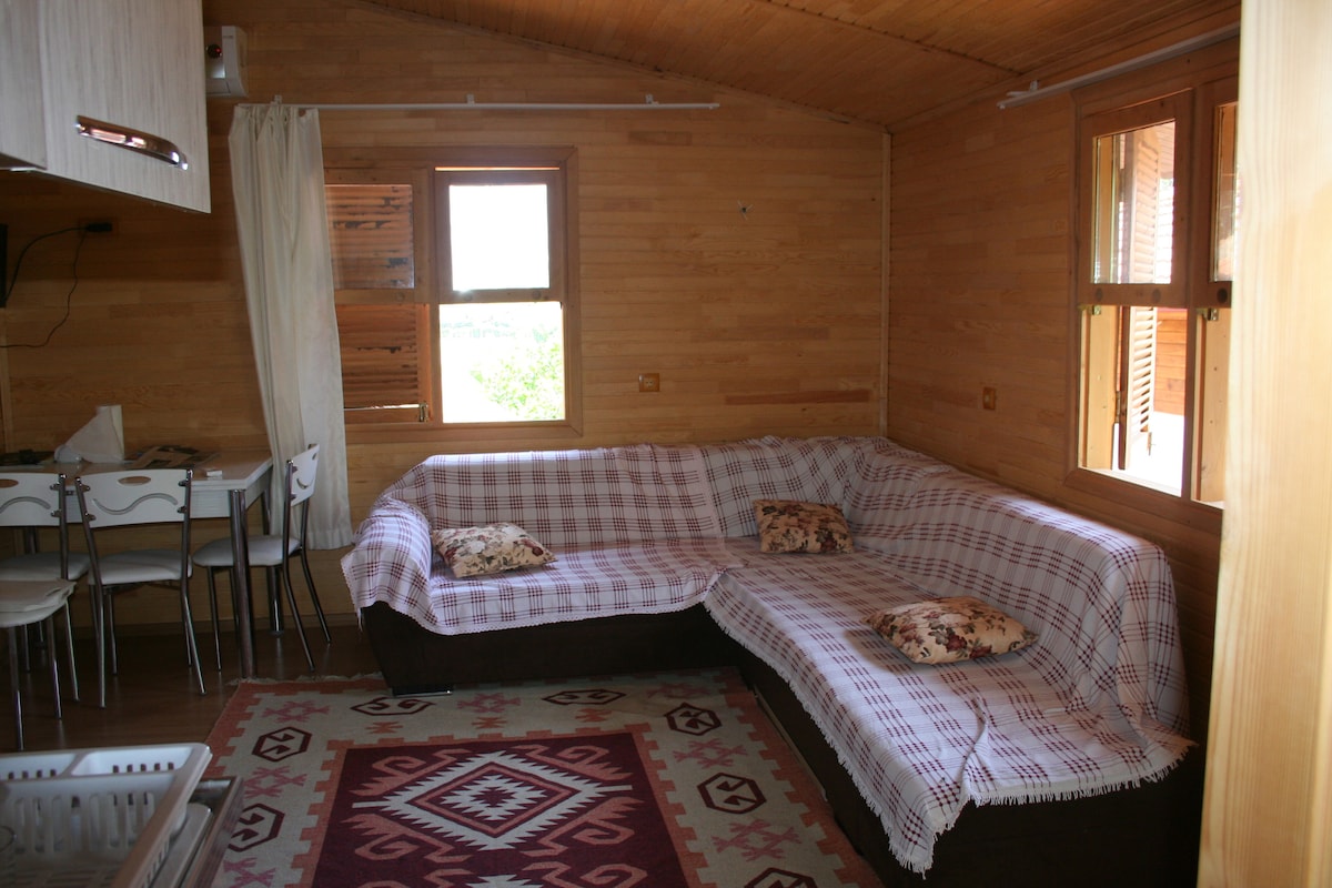 A living area features a spacious corner sofa covered with a patterned throw, complemented by decorative cushions. Above, natural light enters through two wooden-framed windows, while a dining table with white chairs is set against a backdrop of wooden paneling.