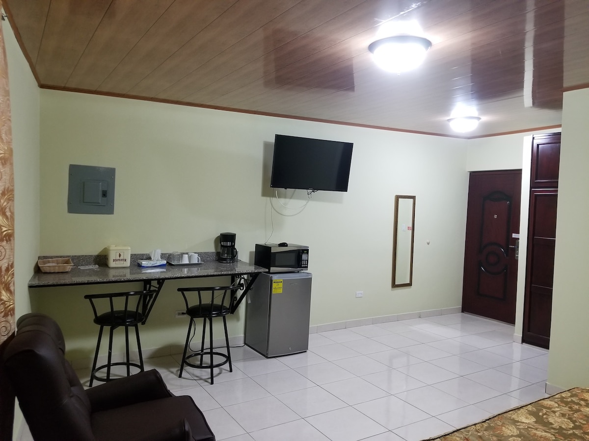 A clean and functional space is shown, featuring a small kitchenette area with a counter and two bar stools. A flat-screen television is mounted on the wall. A microwave and a mini-refrigerator are positioned nearby. Soft lighting enhances the neutral colors of the walls and flooring.