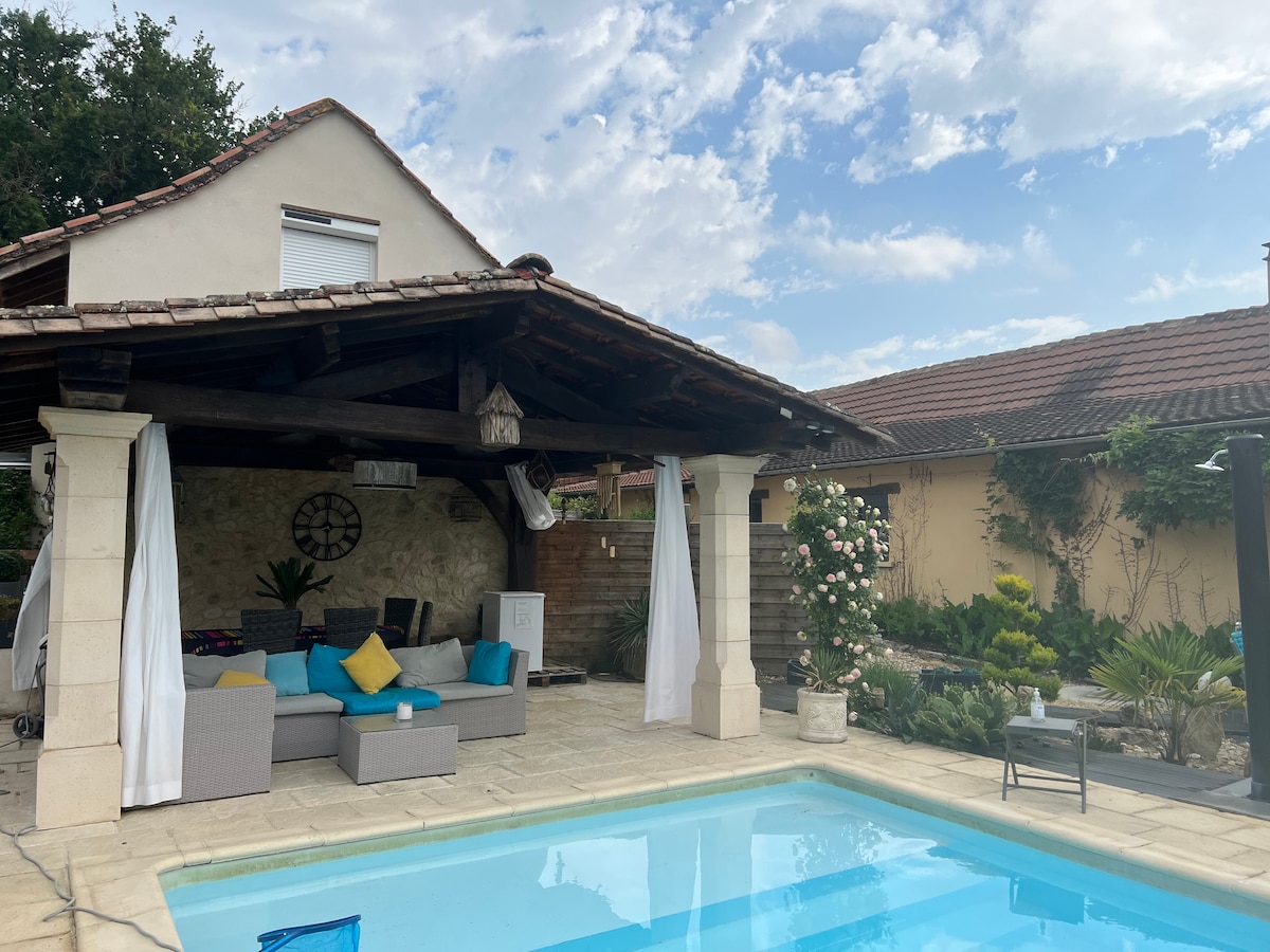 A shaded patio area offers comfortable seating arrangements near the pool, surrounded by lush greenery and decorative plants. The design features soft curtains that allow for light filtering, enhancing the open atmosphere. A well-maintained pool reflects the clear sky above.
