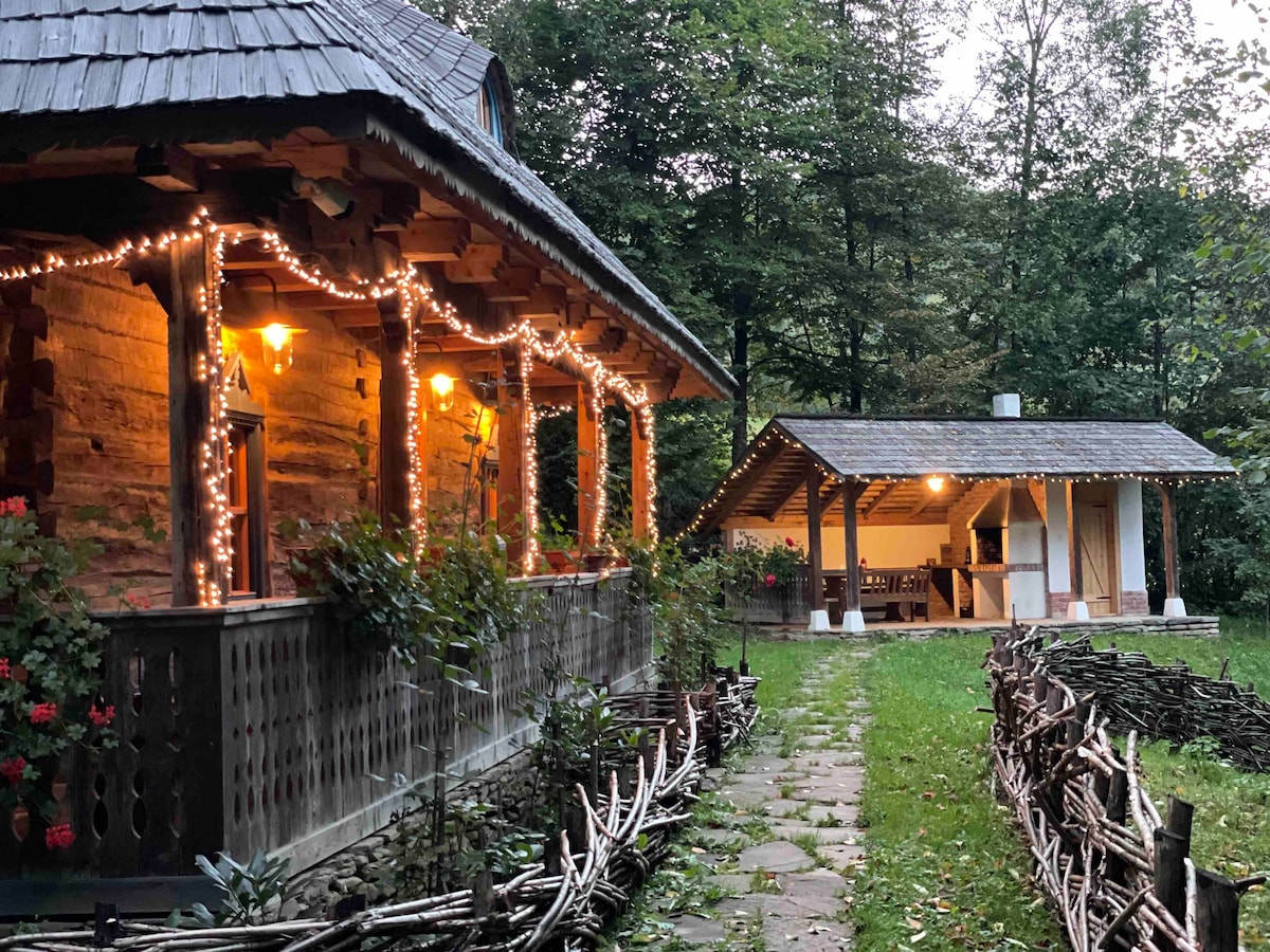 A rustic wooden cabin is illuminated by warm lights, highlighting its porch and surrounding greenery. A stone pathway leads to an open shelter with a cooking area, enhancing the inviting outdoor space. Nature frames the area, creating a peaceful atmosphere.