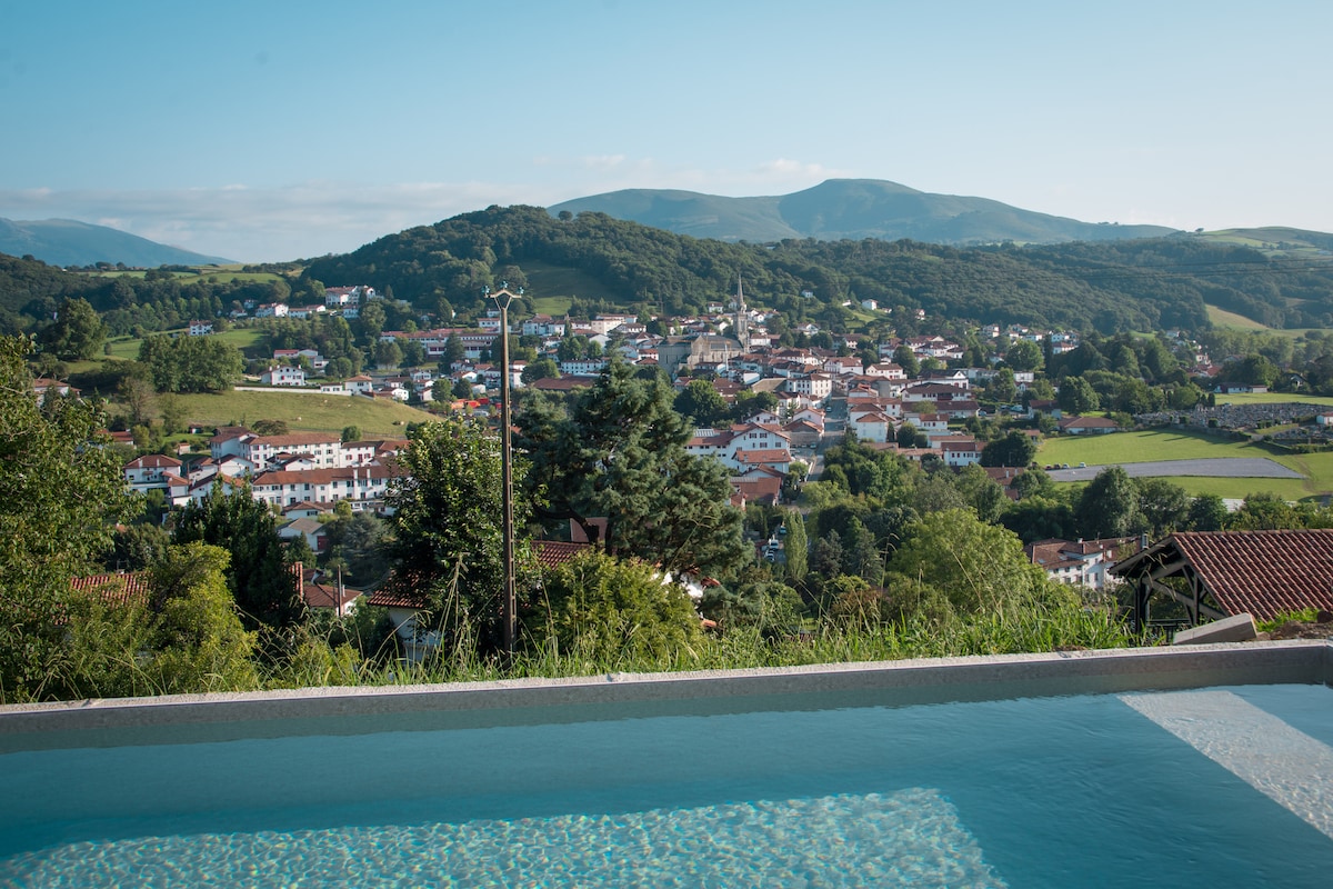 A serene view is offered from an infinity pool, overlooking the village of Hasparren and the surrounding lush greenery. Rolling hills and mountains create a tranquil backdrop, while the pool's clear water reflects the blue sky.