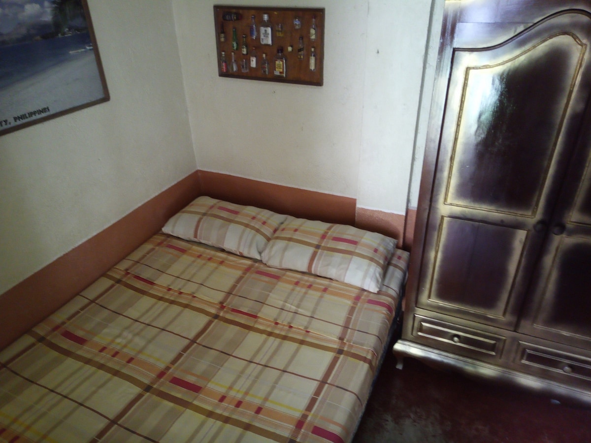 A cozy sleeping area features a queen bed with plaid bedding. A wooden wardrobe stands nearby, providing storage space. The wall displays a bulletin board, adding a personal touch to the room. Natural light enters softly, creating a welcoming atmosphere.