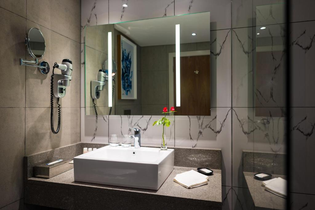 A modern bathroom features a sleek, white basin set on a dark granite countertop. Flanking the basin, two mirrors are illuminated by vertical lighting. A small vase with red flowers adds a touch of color, with amenities neatly arranged nearby.