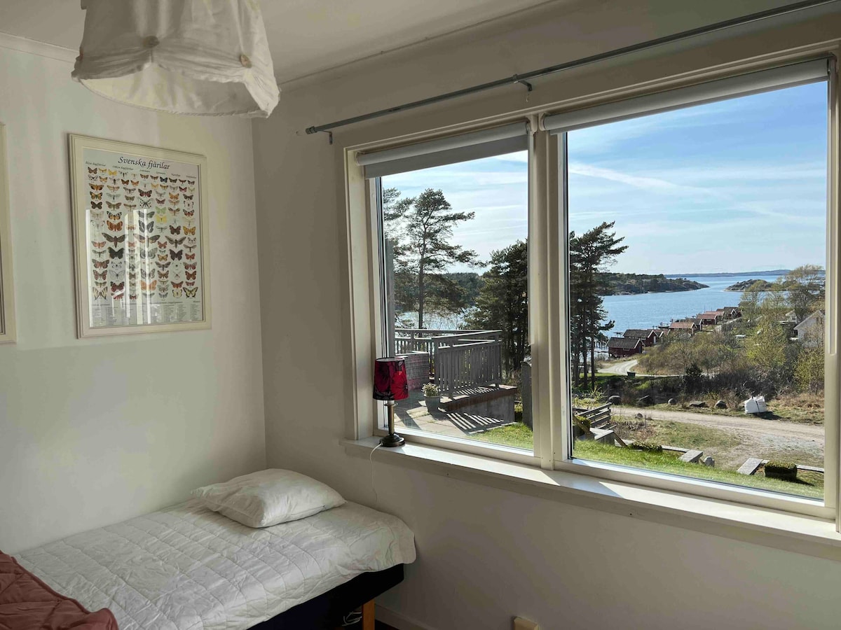 A cozy room features a spacious bed with a soft white blanket. Large windows offer a view of the coastline, capturing the serene water and trees outside. A bedside lamp adds a touch of warmth, and butterfly art is displayed on the wall.