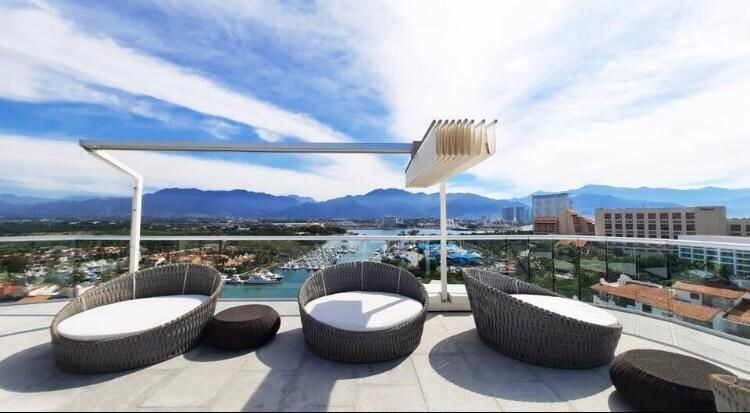 A spacious rooftop lounge area is showcased, featuring modern round furniture with white cushions. The view overlooks a serene marina with boats lined up and mountains in the background. The open sky displays soft clouds, enhancing the feeling of tranquility.
