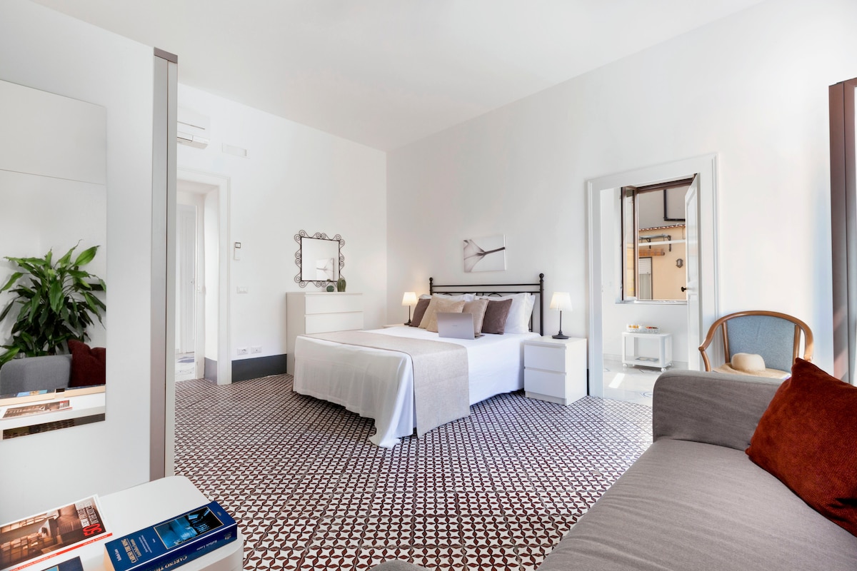 A spacious bedroom is fitted with a large bed, complemented by soft linens and decorative pillows. A gray sofa is positioned nearby, while natural light filters through a window. The patterned floor adds a touch of character, and a subtle plant enhances the ambiance.