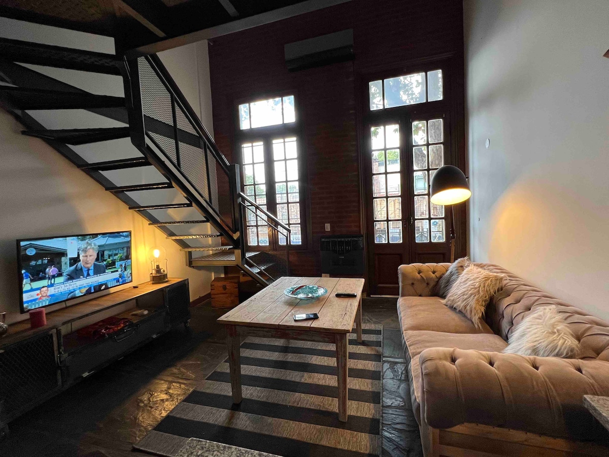 A modern living area is highlighted by large windows allowing natural light. A comfortable sofa faces a television, complemented by a wooden coffee table. A staircase leads to the upper level, and a textured area rug adds a warm touch to the inviting space.