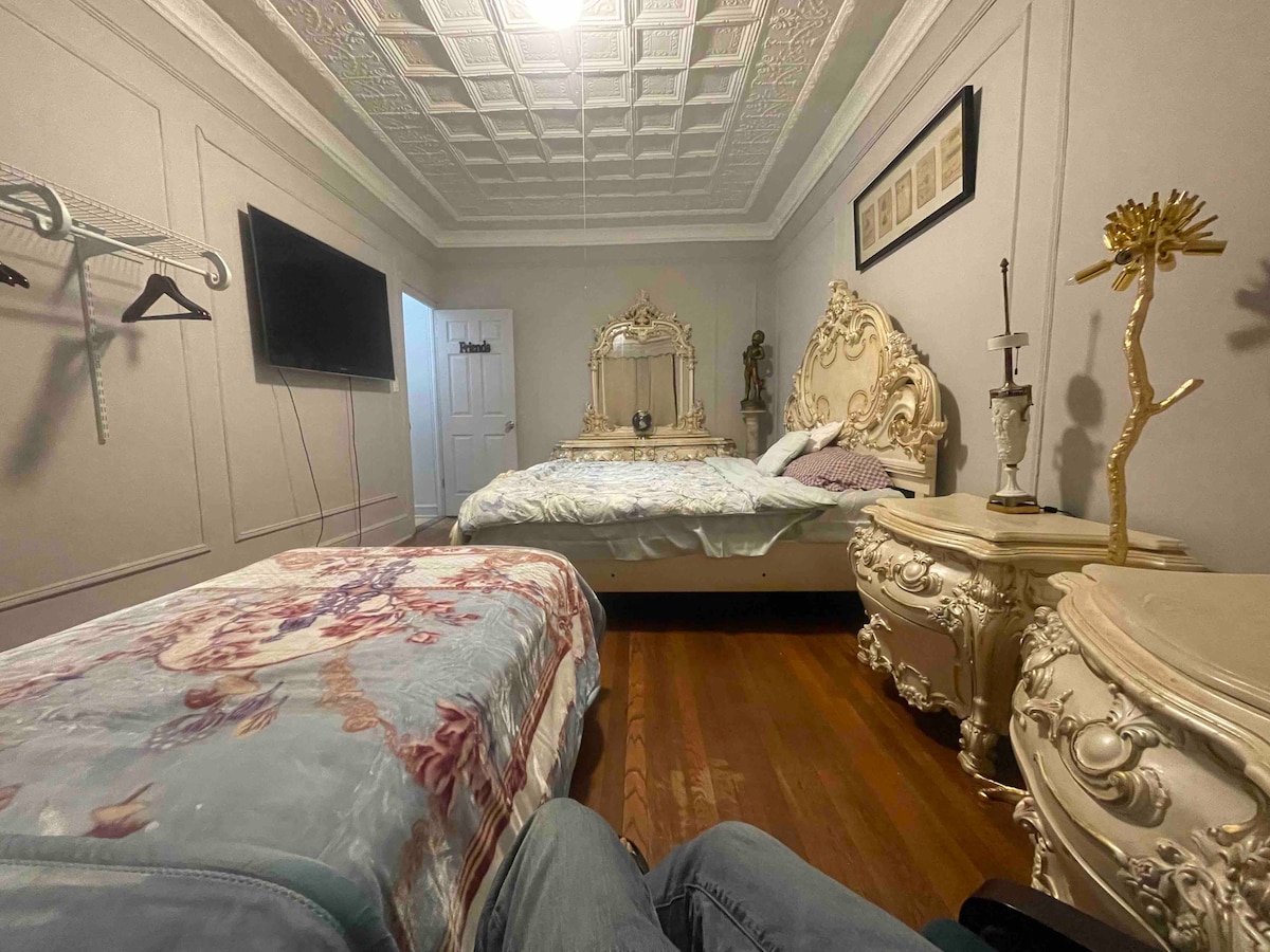 A shared bedroom features ornate decor with two beds positioned opposite each other. Decorative furniture includes a carved dresser and a bedside table with a lamp. A television is mounted on the wall. The flooring is wooden, adding warmth to the space.