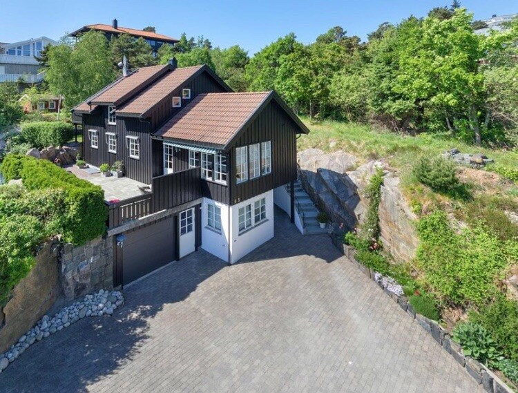 The two-story house with a dark exterior features a sloped roof and multiple windows that allow ample natural light. A spacious driveway made of pavers leads to the entrance, with landscaped gardens and greenery surrounding the property, offering a sense of openness and tranquility.