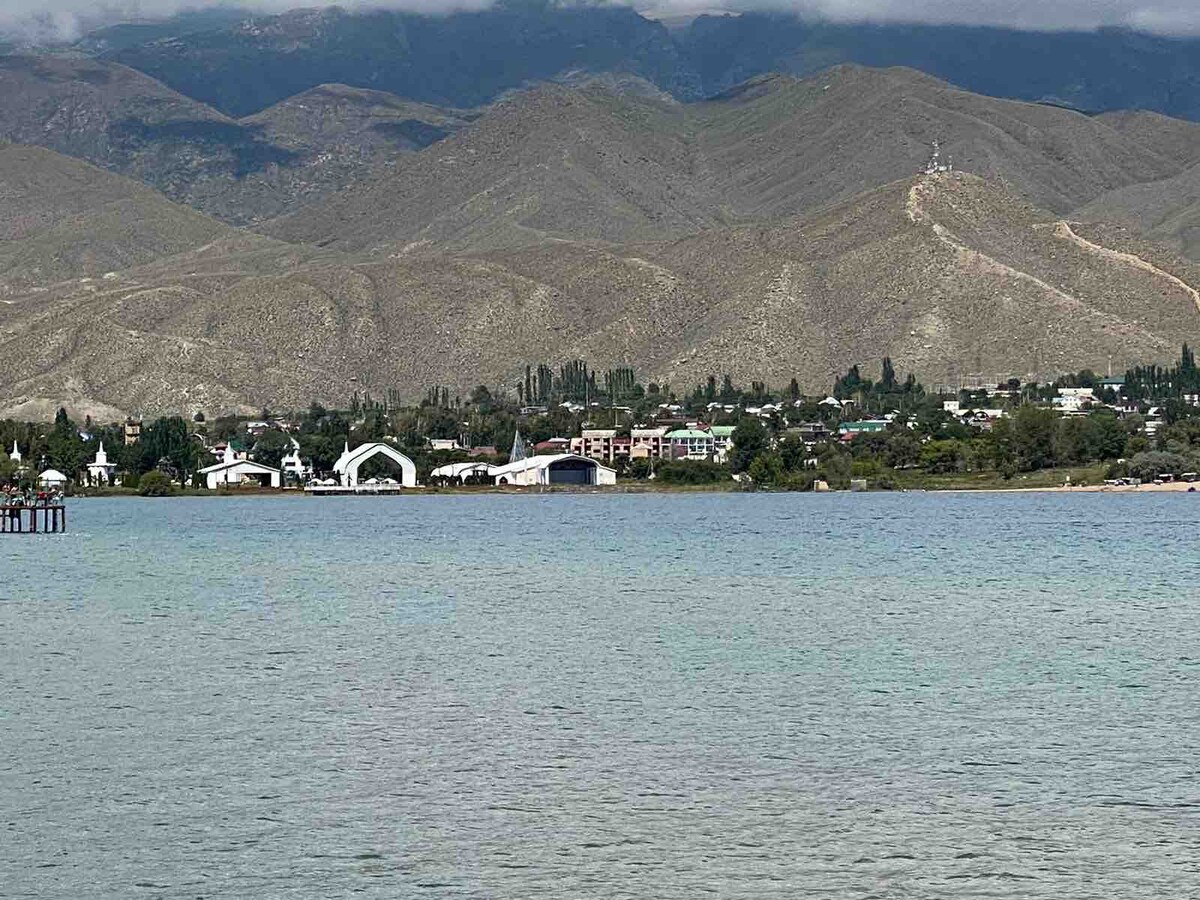 A scenic view of a calm body of water reflects the surrounding mountains and greenery. The distant town features low-rise buildings set against a backdrop of rolling hills, creating a serene and natural atmosphere.