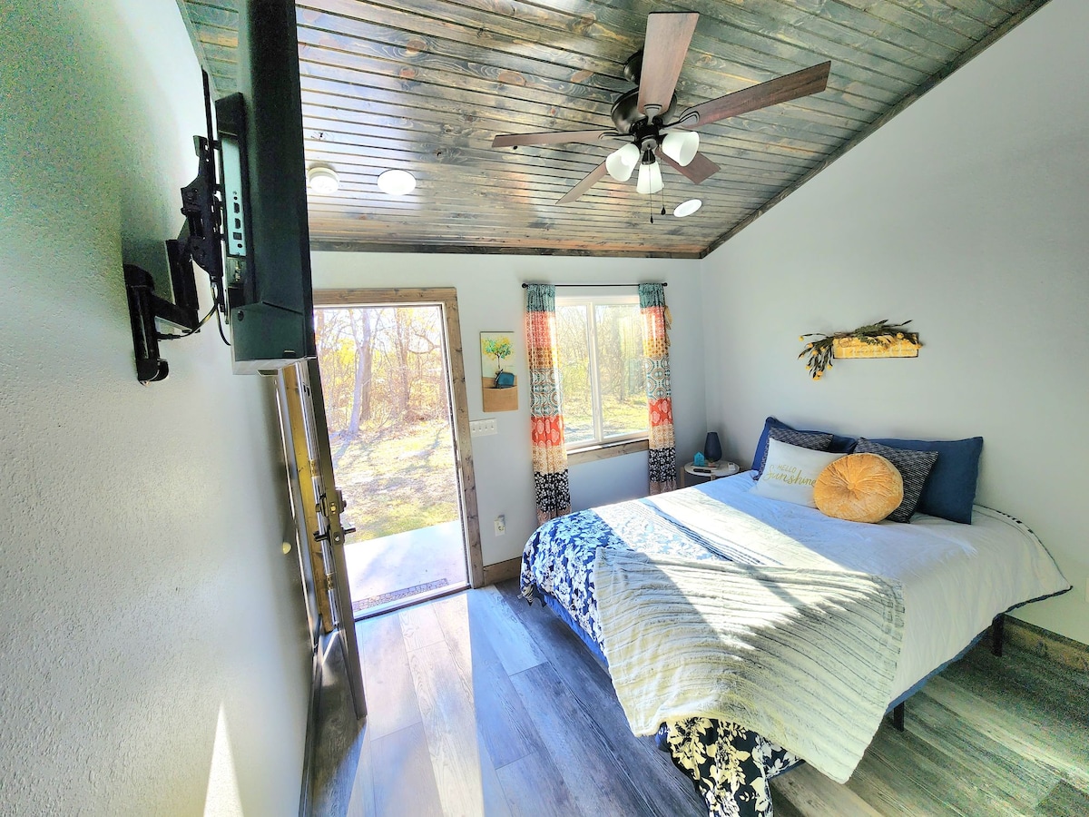 A well-lit room features a spacious bed adorned with decorative pillows. A ceiling fan provides comfort, while natural light enters through a nearby window and door, which opens to an outdoor view. A television is mounted on the wall for entertainment.