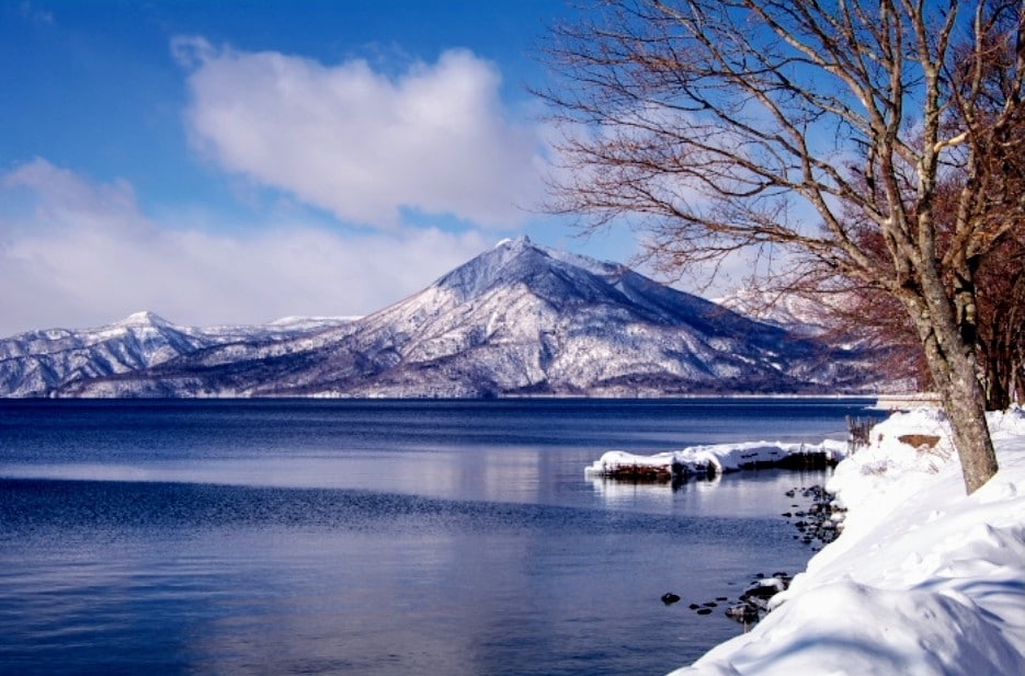 A serene winter scene showcases a lake bordered by snow-covered shores. Majestic mountains are visible in the background, their peaks dusted with snow. The clear sky allows wisps of clouds to drift, creating a tranquil atmosphere.
