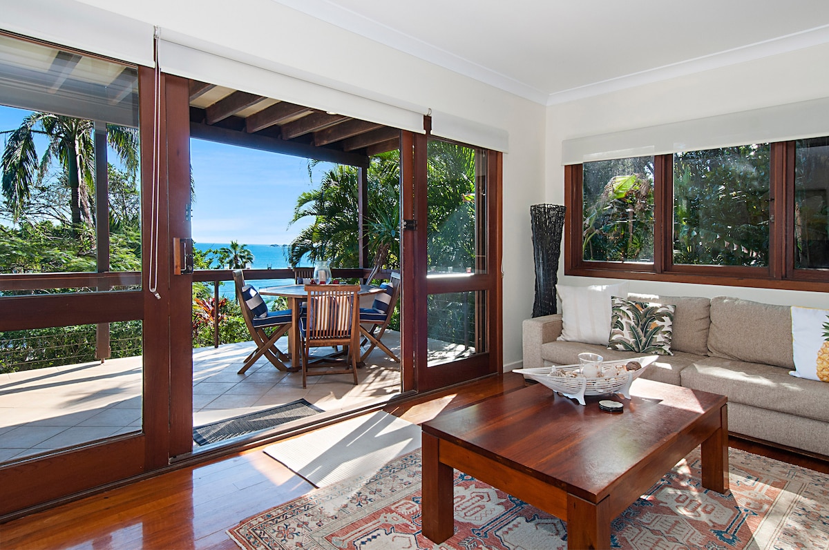 A comfortable living area is framed by large glass doors that open to a patio with ocean views. A wooden coffee table sits atop an intricately patterned rug, while a light-colored sofa provides seating. Lush greenery surrounds the space, enhancing the bright atmosphere.