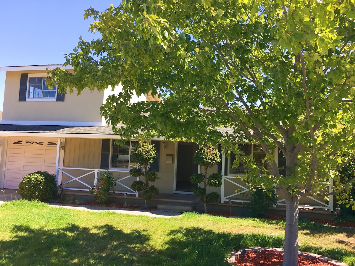 The exterior of a two-story house is shown, framed by a green lawn and decorative shrubs. A large tree provides shade, while a covered porch with white railings welcomes visitors. The garage door is partially visible on the left side.