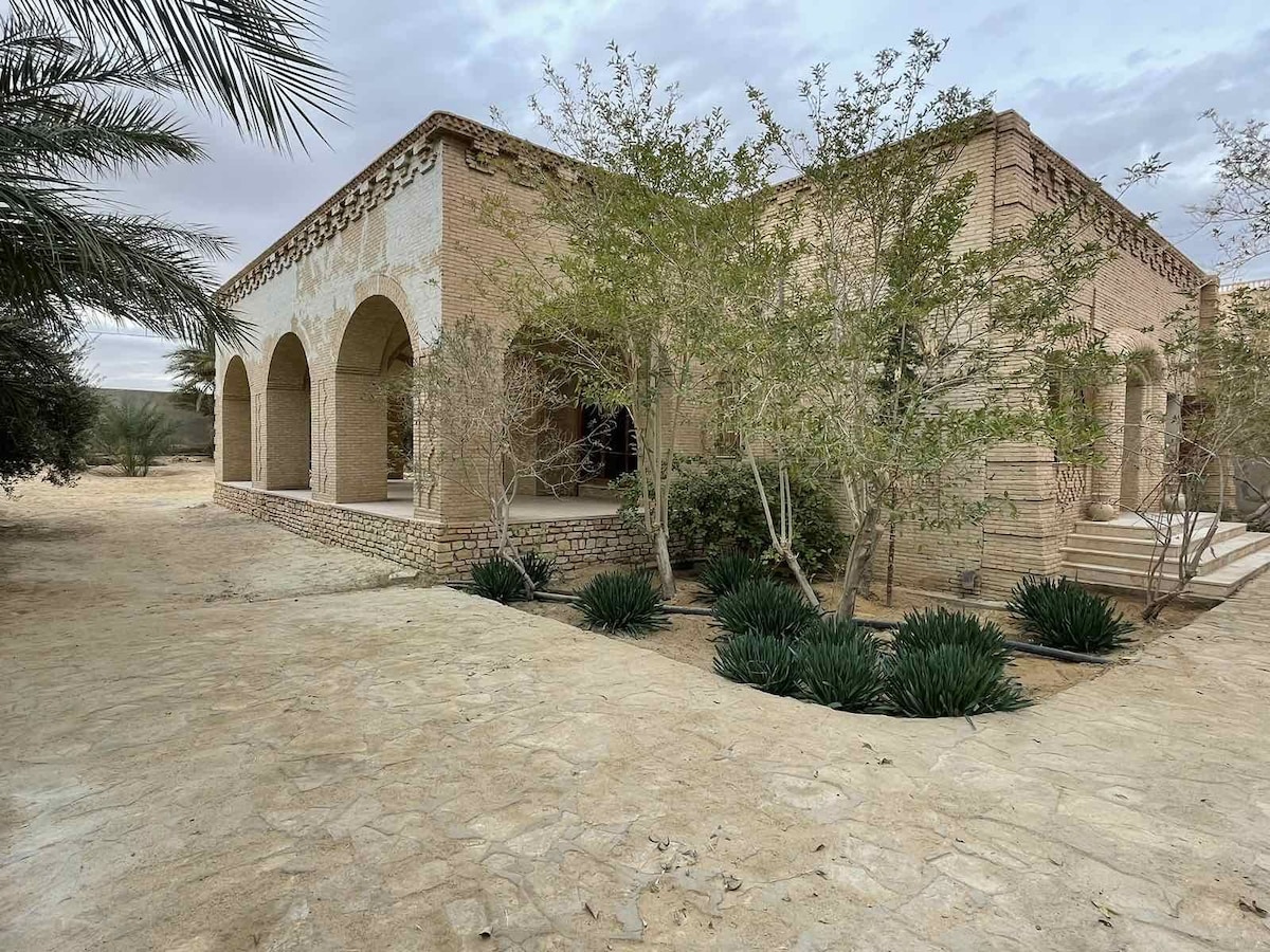 A serene exterior view of a spacious adobe structure is framed by archways. Surrounding greenery includes neatly trimmed shrubs and small trees, enhancing the natural setting. A stone pathway leads toward the entrance, providing a welcoming approach.