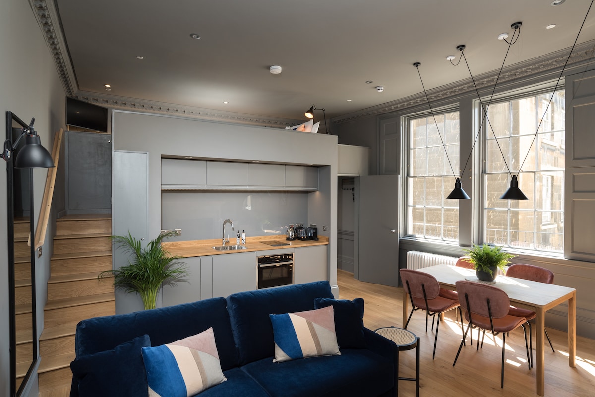 The open-plan living area features a navy blue sofa with decorative cushions, adjacent to a kitchen area with modern appliances. A dining table with pink chairs is set against large windows, allowing ample natural light to fill the space. Wooden stairs lead to the mezzanine level.