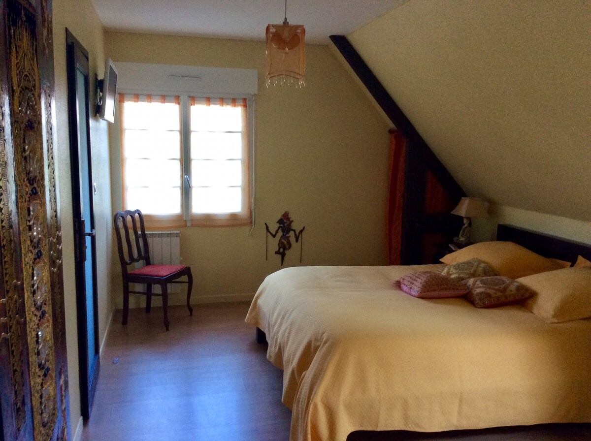 A bright bedroom features a spacious bed covered in soft yellow linens, complemented by decorative pillows. Natural light enters through a window with wooden shutters. A vintage chair is positioned nearby, alongside a small bedside lamp. Decorative wall art adds character to the room.