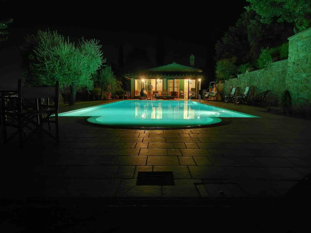 A serene outdoor pool is highlighted by soft turquoise lighting, set against a natural stone backdrop. In the distance, a charming gazebo and lounge chairs are visible, surrounded by greenery, while trees and illuminated spaces create a peaceful evening ambiance.