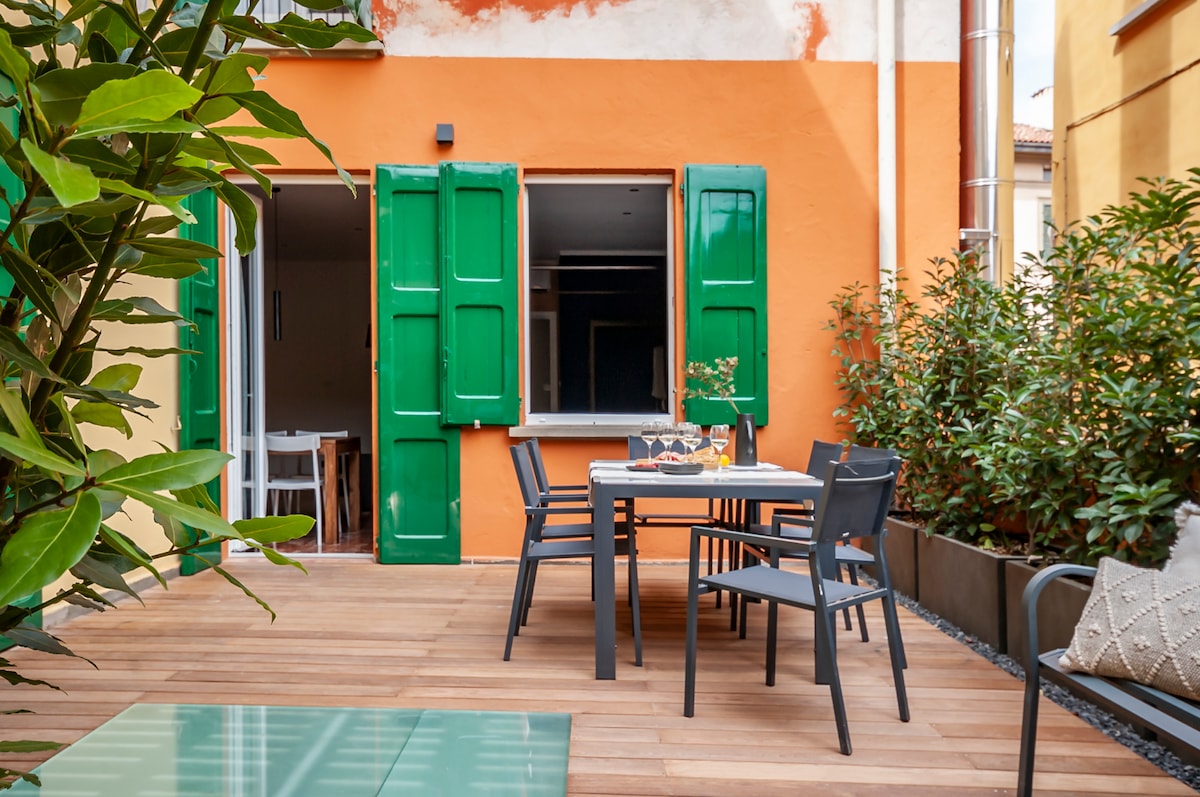 An outdoor terrace features a wooden deck surrounded by greenery. A dining table with chairs is set for an inviting meal, with large green shutters framing the view of the interior. Potted plants enhance the tranquil ambiance of the space.