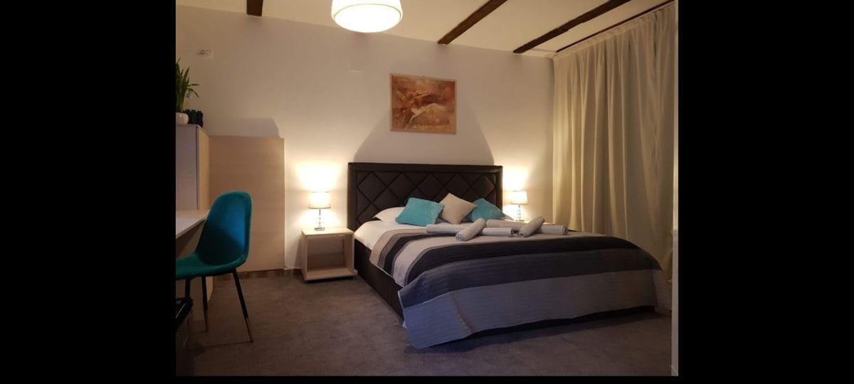 A spacious bedroom features a large bed with a patterned blanket and several pillows. Soft lighting is provided by bedside lamps on each side. A modern chair and a small desk are positioned nearby, and sheer curtains frame a window, adding to the serene ambiance.