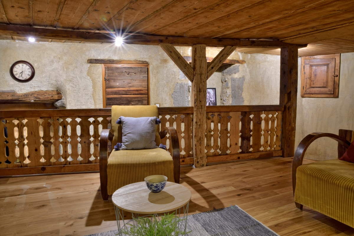 A warm wooden interior features exposed beams and rustic touches. A cozy armchair and a yellow striped chair are positioned around a small round table. The space is illuminated by soft lighting, highlighting the charm of natural materials and textures.