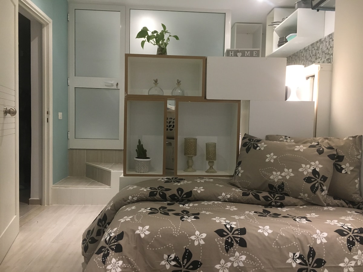 A compact studio features a neatly arranged sleeping area with a floral-patterned bedding set. A decorative partition separates the space, showcasing small plants and tasteful accents. A soft light is provided by fixtures, highlighting a calm and functional atmosphere.