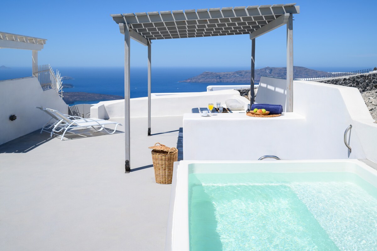 A private terrace features a hot tub and lounge chairs, set against a backdrop of expansive views of the Aegean Sea. A small table with refreshments is positioned nearby, enhancing the serene atmosphere of the space.
