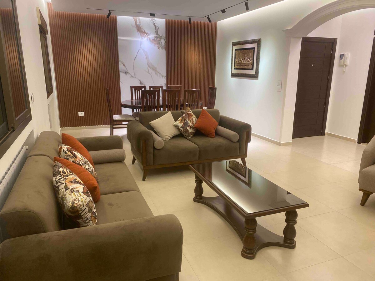 A sophisticated living area is furnished with a large brown sofa adorned with decorative cushions. A sleek coffee table is placed at the center, and a dining table with wooden chairs is visible in the background. Soft lighting enhances the warm atmosphere of the space.