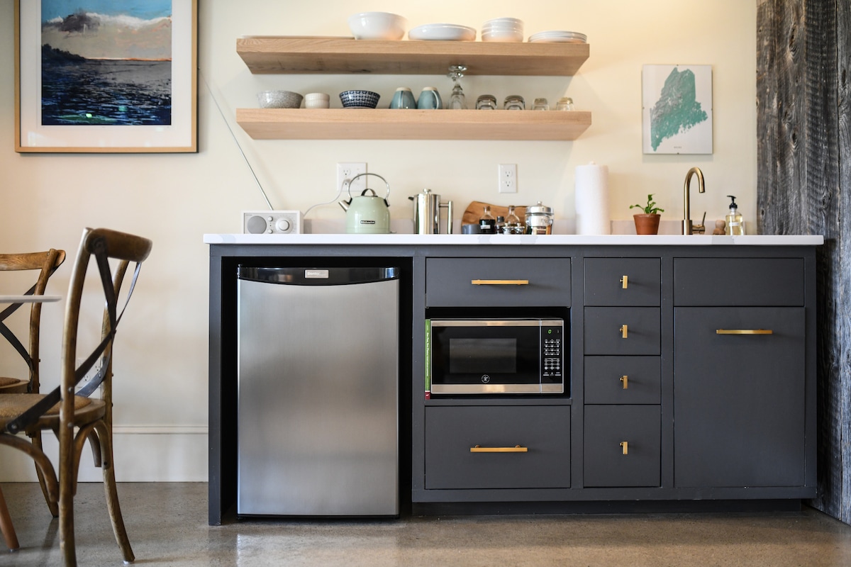 The kitchenette features sleek, dark cabinetry with gold hardware, a stainless steel refrigerator, and a microwave. Open shelving displays various ceramic dishes and glassware. A small sink with a plant beside it adds a touch of greenery, while a cozy dining area with wooden chairs is nearby.