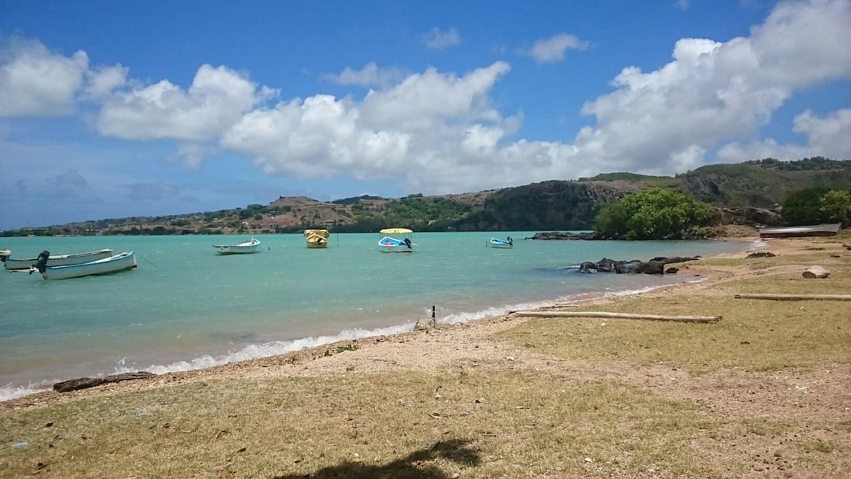 A serene beach scene features calm turquoise waters with several small boats anchored close to the shore. Lush green hills rise in the background under a partly cloudy blue sky, while the sandy beach is lined with gentle waves and scattered rocks.