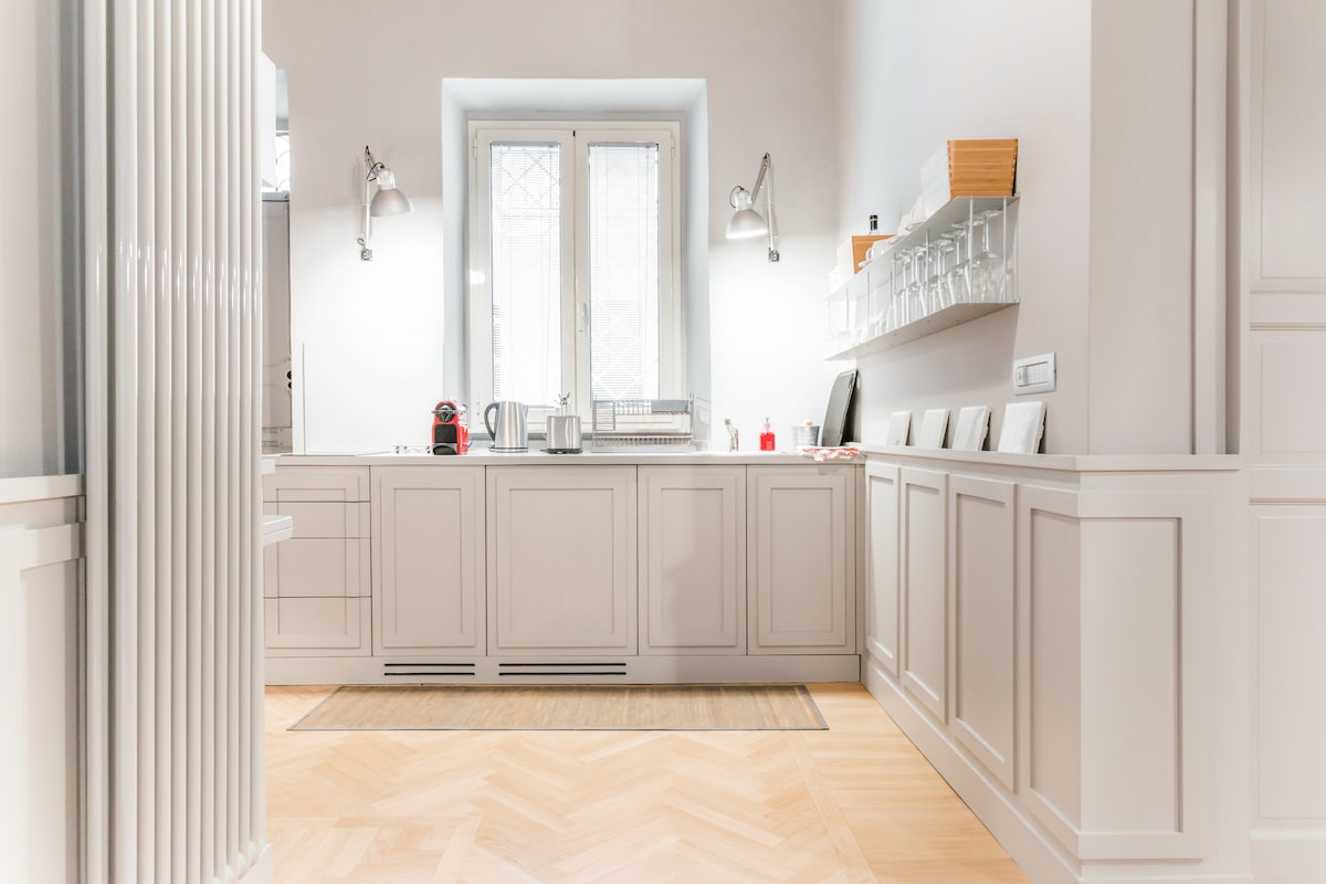 A modern kitchen is presented with light cabinetry and a spacious layout. Natural light illuminates the room through two tall windows, while various kitchenware is neatly arranged on open shelves. A runner rug sits on the light hardwood floor, adding a touch of warmth.