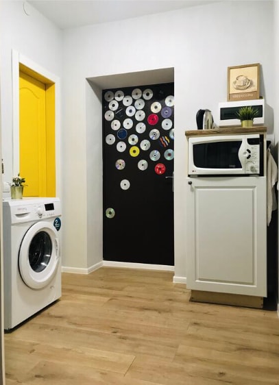 A functional laundry corner is displayed, featuring a washing machine and a white microwave against a backdrop of playful wall decor made from circular CDs. A bright yellow door adds a pop of color to the neutral-toned walls and natural wood flooring.