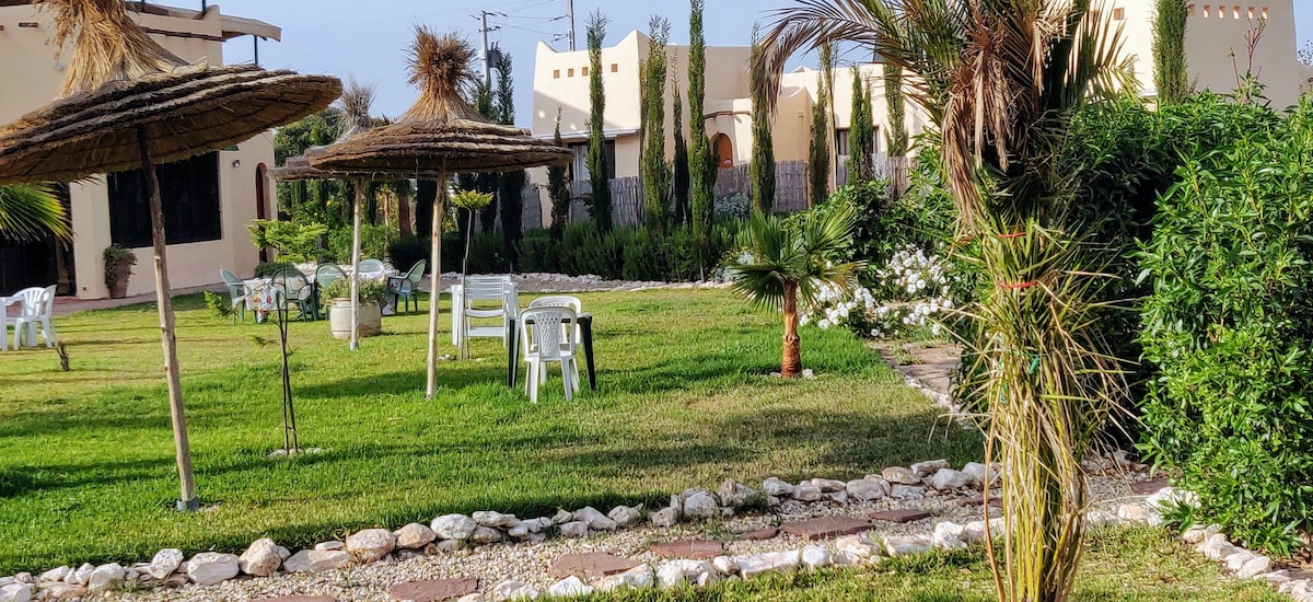 A landscaped garden area features multiple shaded seating arrangements under straw umbrellas. Lush greenery and decorative plants surround the space, with a pathway made of stones guiding through the area. White flowers add a touch of color to the serene outdoor setting.