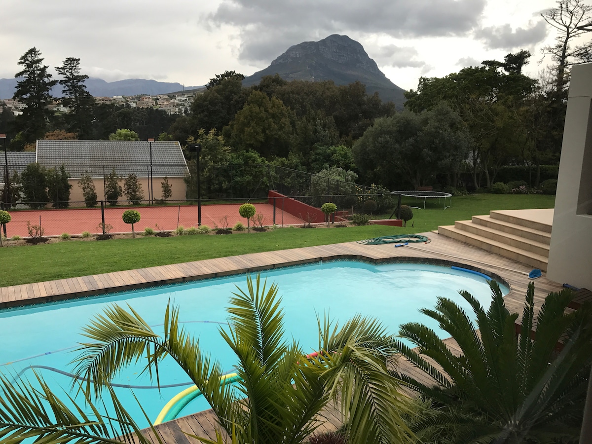 A view of a large swimming pool surrounded by wooden decking and lush greenery, with a tennis court visible in the background. The backdrop features a mountainous landscape under a soft, cloudy sky.