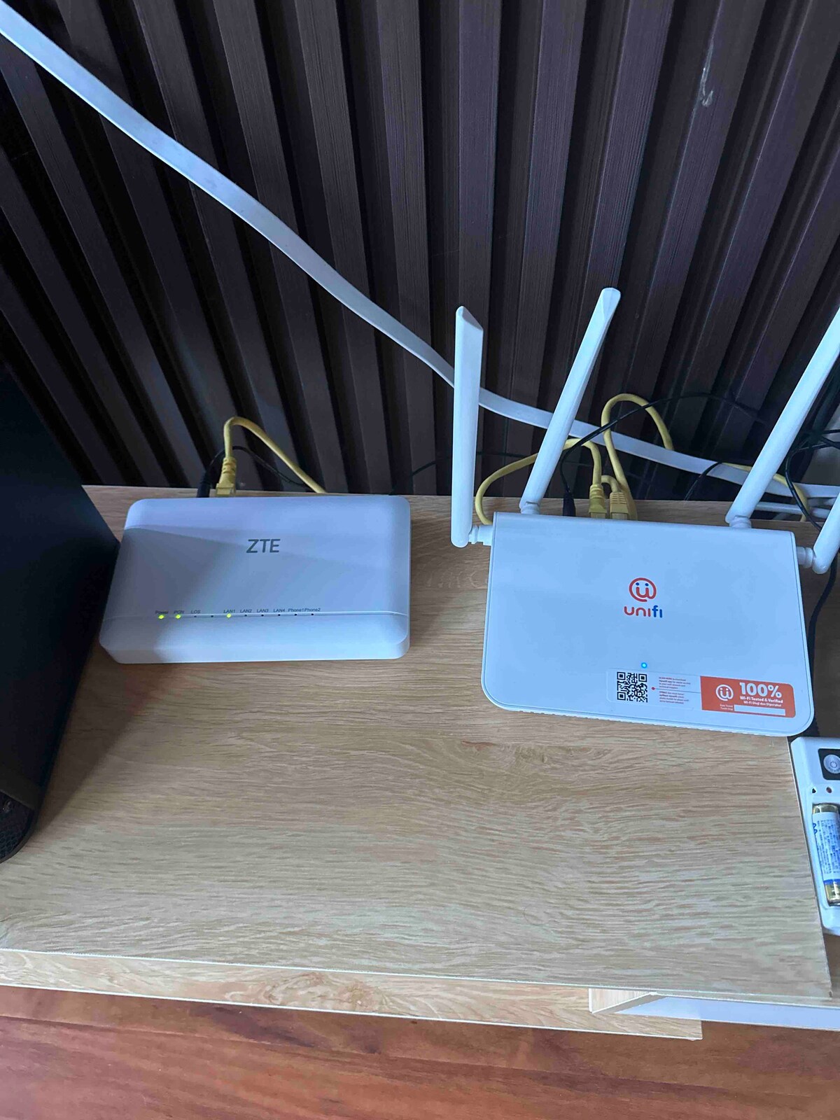 Two internet routers are placed on a wooden surface, with visible LED indicators showing connectivity status. The routers feature distinct designs, one being white with a logo and the other silver. Behind them, cables are neatly organized against a brown paneling.