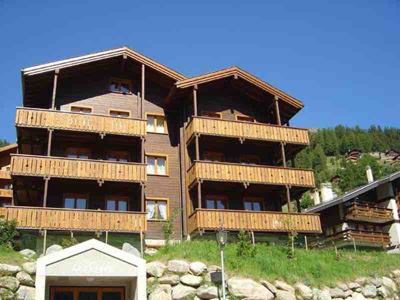 A three-story Swiss chalet features wooden balconies with railing on each level. Large windows provide ample natural light. The building is surrounded by a grassy area and small rocks, set against a backdrop of green hills and a clear blue sky.