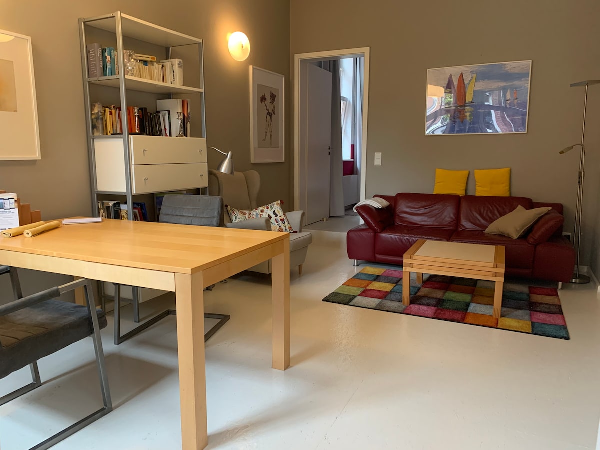 A combined living and dining area features a light wooden table accompanied by four chairs. A red sofa is paired with vibrant yellow cushions, and a colorful rug lies beneath a coffee table. A bookshelf stands against the wall, filled with various books.