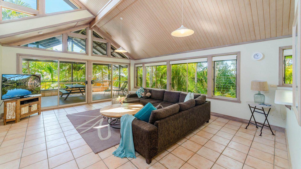 A spacious living area features a large dark sectional sofa with turquoise accents, situated on tiled flooring. Expansive windows provide abundant natural light and views of the surrounding greenery. A circular coffee table is centered on a soft area rug, with a small side table nearby.