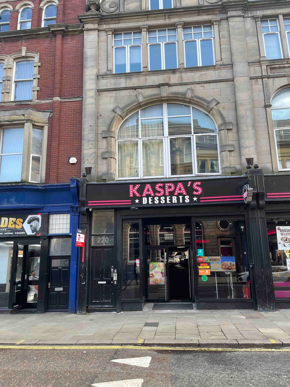 A storefront features the sign for Kaspa's Desserts prominently displayed in pink lettering. The entrance is framed by large windows and ornate architectural details, while neighboring shops contribute to the vibrant urban atmosphere.