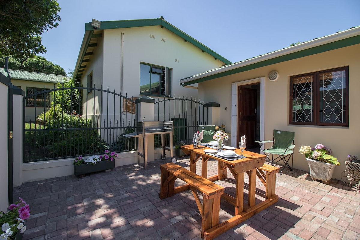 An outdoor dining area features a wooden table with seating, set against a backdrop of a charming home. Lush greenery and colorful flowers enhance the inviting atmosphere. A barbecue grill is positioned nearby, and light filters through the clear sky above the fenced space.