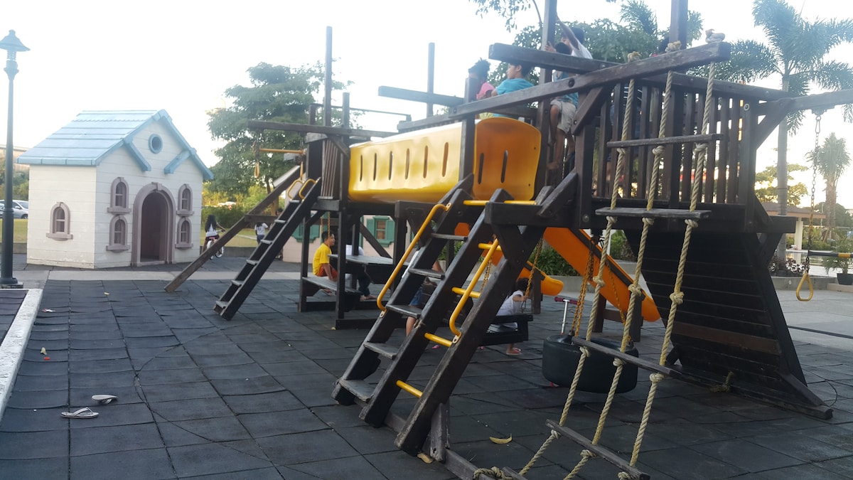 A colorful playground features a large wooden structure with slides, climbing ropes, and ladders. Adjacent stands a small playhouse with a peaked roof and decorative windows, surrounded by safety mats. Lush greenery serves as the backdrop, creating an inviting outdoor play area.
