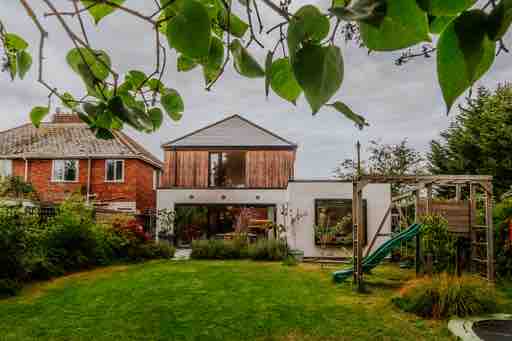 A modern two-story house is set within a spacious yard, featuring a mix of wooden and white exterior walls. Lush greenery surrounds the property, and outdoor play equipment is visible, including a slide. Large windows reflect the surrounding nature, creating a harmonious outdoor space.