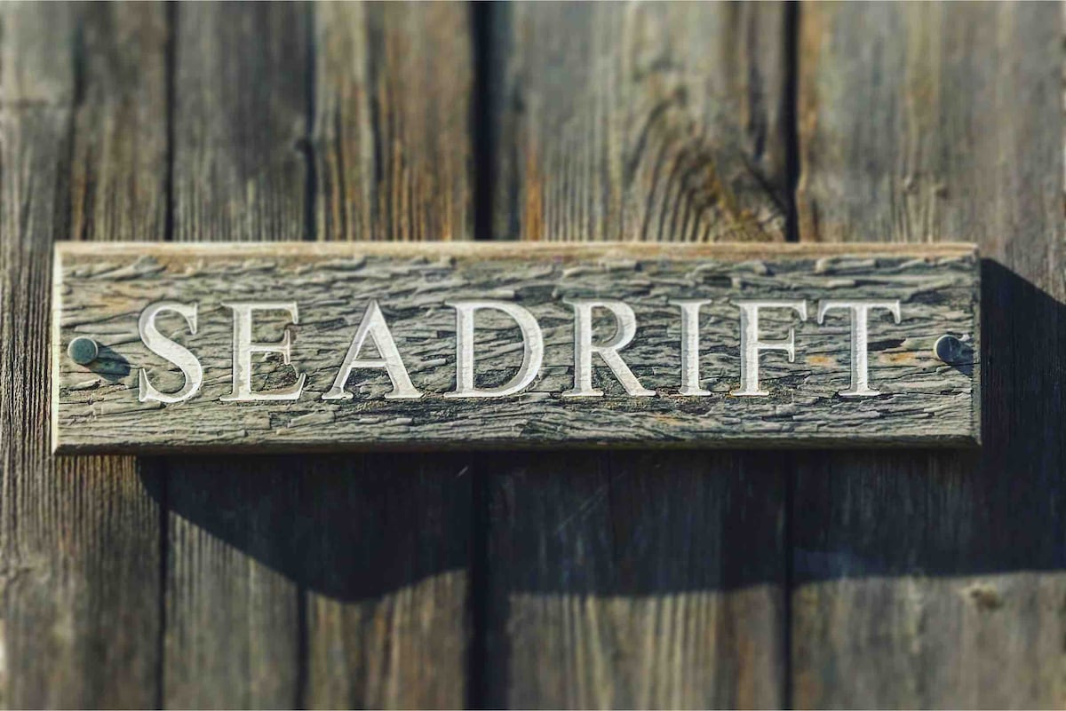 The wooden sign displaying the name 'SEADrift' is mounted on a textured, weathered wood background. The natural tones of the wood complement the rustic aesthetic, with shadows cast by the lettering creating subtle contrasts.