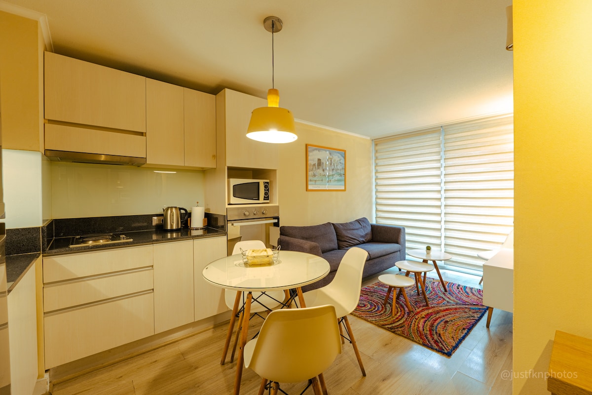 A modern kitchen space is seen with sleek cabinetry and a compact dining table surrounded by four chairs. A comfortable sofa sits in the corner, complemented by a vibrant area rug. Natural light filters through the partially open blinds, creating a bright atmosphere.