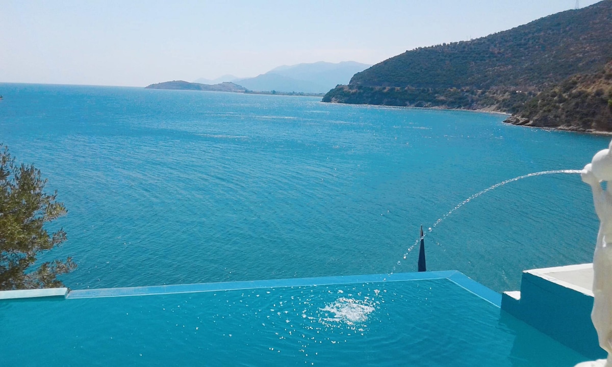 A tranquil scene features a vibrant blue sea, extending towards distant hills and a clear sky. A sculpted fountain gracefully adds to the view, with water gently flowing into a plunge pool that complements the serene atmosphere.