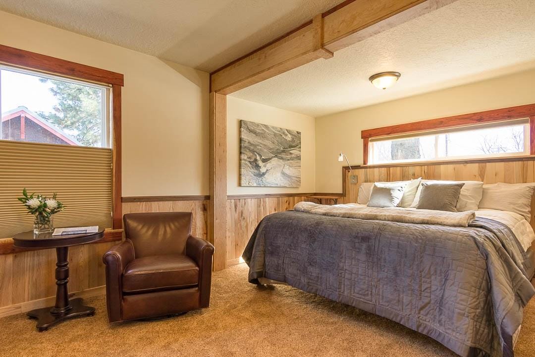 A spacious bedroom features a luxurious bed dressed in soft linens, complemented by two windows allowing natural light. A comfortable armchair is positioned next to a side table, which holds a small vase with fresh flowers. Wood paneling adds warmth to the room.